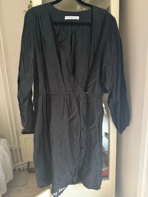 Svart omlottklänning Rodebjer  - Svart omlottklänning från Rodebjer med långa ärmar och fina spetsdetaljer längs omlottkanten. Klänningen har en avslappnad passform och är perfekt för dig som gillar stilrena plagg med en twist.