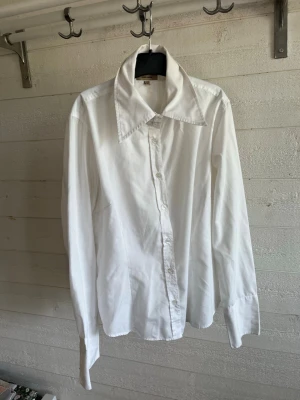 Vit skjorta från Vero Moda - Klassisk vit skjorta från Vero Moda med lång ärm och stora spetsiga kragar. Skjortan har knäppning framtill och är tillverkad i mjuk bomull. Perfekt att styla till jeans eller kjol för en clean look.