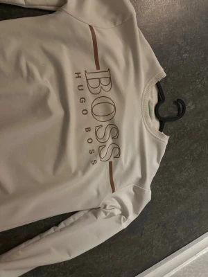 Vit sweatshirt från Hugo Boss - Snygg vit sweatshirt från Hugo Boss med stort bronze BOSS-tryck och ribbad rund hals. Tröjan har långa ärmar och en clean, stilren look som passar perfekt till jeans eller joggers.