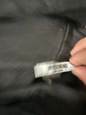 Svart Moschino hoodie med björntryck - Snygg svart hoodie från Moschino med stort tryck framtill. På bröstet står det 'Moschino Couture!' i vitt och guld, och under syns en söt björn i skinnjacka och keps. Hoodien har huva med dragsko och är tillverkad i mjukt material.