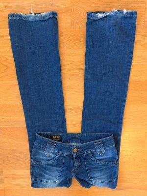 Blå Lee bootcut jeans - Säljer ett par klassiska blå bootcut jeans från Lee med snygga detaljer på fickorna och kontrastsömmar. Jeansen har fem fickor, knappgylf och Lee-logga bak i midjan. Perfekta för dig som gillar en tidlös och avslappnad stil.