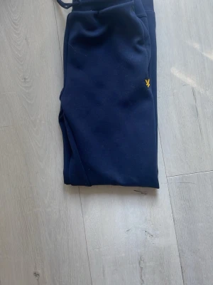 Mörkblå mjukisbyxor med broderad logga - Snygga mörkblå mjukisbyxor med dragsko i midjan och resår vid bensluten. Byxorna har en gul broderad logga på ena benet och är tillverkade i ett mjukt och bekvämt material. Perfekta för chill eller träning.