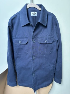  Wahlberg mörkblå overshirt - Stilren mörkblå overshirt med klassisk krage och fyra knappar framtill. Två bröstfickor med lock och knapp samt diskreta sidofickor. Storlek M. Jag har bara haft den på mig 1 gång men märkte att den var för tajt på mig (187 cm 82kg). Perfekt skick. Ny pris 1600kr