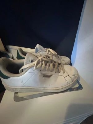 Vita Adidas Stan Smith sneakers - Klassiska vita Adidas Stan Smith sneakers med gröna detaljer på hälen. Skorna har lågt skaft, vita snören och diskret Adidas-logga på sidan. Tillverkade i slätt läder med enkel och stilren design som passar till det mesta.