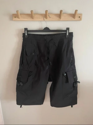 Svarta cargoshorts med snörning - Säljer ett par svarta cargoshorts med flera praktiska fickor och snörning i midjan. Shortsen har en loose passform och är perfekta för dig som gillar streetwear eller outdoor-vibe. Tillverkade i ett lätt syntetmaterial som känns skönt under sommaren. Passar mig som brukar ha storlek 44 eller 28/29. Använda vid ett tillfälle, som nya.