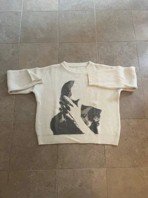 Beige stickad tröja med tryck - Säljer en beige stickad tröja med ett stort grafiskt tryck i grått och svart framtill. Tröjan har rund halsringning och långa ärmar. Materialet ser ut att vara mjukt och bekvämt, perfekt för kyligare dagar. Passar dig som gillar unika plagg med coola motiv.