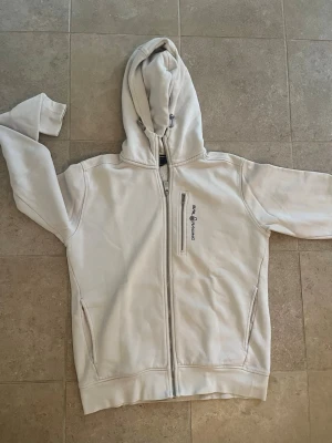 Vit hoodie från Sail Racing - Säljer en vit hoodie från Sail Racing i storlek M. Hoodien har dragkedja framtill, två sidofickor och en bröstficka med dragkedja. Märkets logga är broderad på bröstet. Tillverkad i mjukt bomullsmaterial och har justerbar huva med snören.