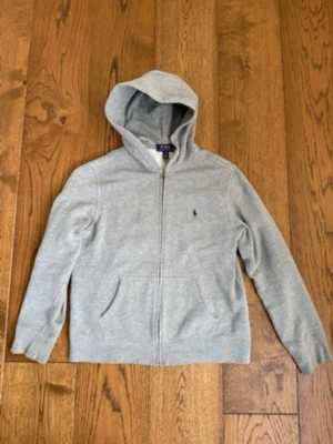Grå hoodie från Polo Ralph Lauren 14-16 år - Snygg grå hoodie från Polo Ralph Lauren med dragkedja framtill och klassisk blå logga broderad på bröstet. Tillverkad i mjukt bomullsmaterial och har två fickor samt en skön huva. Perfekt för en chill och avslappnad stil.