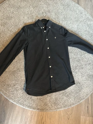 Svart skjorta från Morris - Klassisk svart skjorta från Morris med button-down krage och vita knappar. Skjortan har lång ärm och en broderad logga på bröstet. Tillverkad i mjuk bomull som känns skön mot huden. Perfekt för dig som gillar stilrena plagg med snygga detaljer.