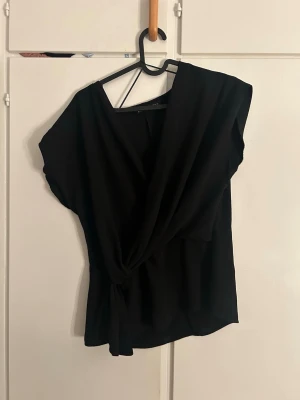 Svart omlottopp från Zara - Säljer en svart omlottopp från Zara i storlek S. Toppen har en snygg omlottdesign med knyt i sidan och korta ärmar. Perfekt till jeans eller kjol för en trendig look.