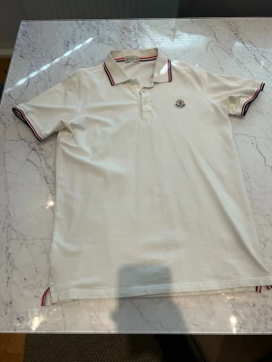 Moncler pike - Klassisk vit pikétröja med korta ärmar och krage. Tröjan har rödblå ränder på krage och ärmslut samt en liten broderad logga på bröstet. Perfekt till en avslappnad men stilren look.