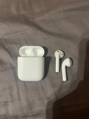 Airpods gen1  - Säljer ett par stilrena trådlösa in-ear hörlurar i vit plast med tillhörande laddningsetui. Hörlurarna har en smidig och kompakt design som gör dem enkla att ta med sig överallt. Perfekta för musik, poddar eller samtal på språng.