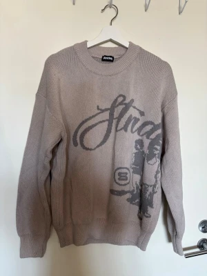 Beige stickad tröja med tryck Strides - Säljer en beige stickad tröja med rund hals. Tröjan har avslappnad passform, långa ärmar och ribbade muddar vid ärmslut och nederkant.