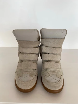 Beige höga sneakers med kardborre - Snygga höga sneakers i beige mocka och textil med tre breda kardborreband och ljusbrun sula. Skorna har perforerade detaljer på tån och en chunky siluett som ger en cool streetwear-vibe.