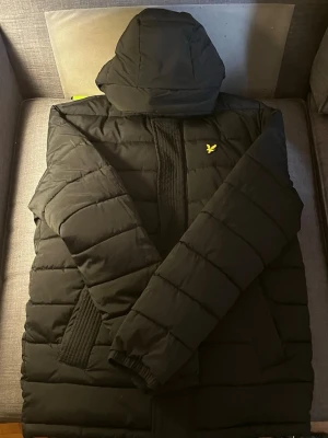 Svart pufferjacka från Lyle & Scott - Säljer en svart pufferjacka från Lyle & Scott med huva och gul logga på bröstet. Jackan har dragkedja framtill, ribbade muddar vid ärmslut och två sidofickor. Perfekt för kalla vinterdagar och riktigt skön att bära. Passar M också