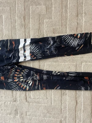 Mönstrade leggings från Stronger XS - Snygga svarta leggings från Stronger i storlek XS med färgglatt abstrakt mönster och vita ränder på ena benet. Tillverkade i 85% återvunnen polyester och 15% elastan för en stretchig och bekväm passform. Perfekta för träning eller chill.