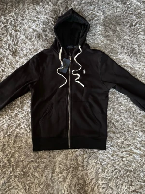 Svart hoodie från Polo Ralph Lauren - Snygg svart hoodie från Polo Ralph Lauren med dragkedja framtill, vit broderad logga på bröstet och vita snören i huvan. Tillverkad i mjukt bomullsmaterial och har två fickor framtill. Perfekt för en avslappnad och stilren look.