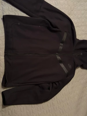 Svart hoodie med dragkedja från Nike - Säljer en svart hoodie från Nike med hel dragkedja framtill och två snedställda fickor. Hoodien har diskreta svarta detaljer på bröstet och en huva. Perfekt för en sportig och avslappnad stil.