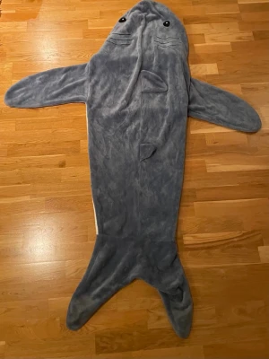 Hajformad sovpåse i fleece - Supermysig sovpåse i form av en haj, perfekt för slappa kvällar hemma. Utsidan är grå med fenor och ögon, insidan är vit. Tillverkad i mjuk fleece som håller dig varm och bekväm. Passar perfekt för att krypa ner i soffan eller sängen.