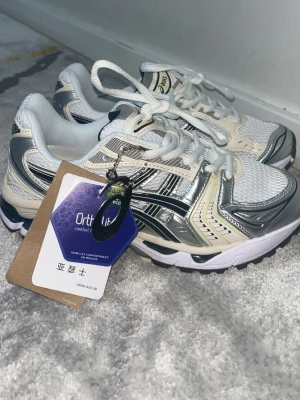 Asics vita och silver sneakers - Snygga Asics sneakers i vitt och silver med mesh- och syntetpaneler. Skorna har klassisk snörning, Ortholite-innersula och retroinspirerad design. Perfekta för dig som gillar sportig stil och bekväma skor.