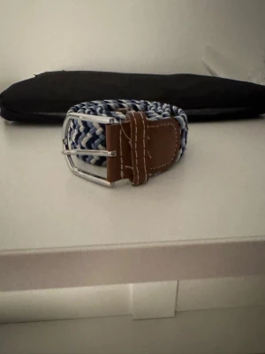 Flätat bälte - Snyggt flätat bälte med randigt mönster i blått och vitt samt detaljer i brunt skinn. 95cm.