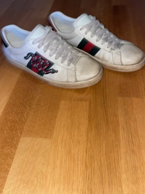 Vita sneakers med orm och ränder - Snygga vita sneakers i läder med klassiska gröna och röda ränder på sidan samt en cool orm-detalj. Skorna har lågt skaft och vita snören. Yttersulan har ett unikt mönster. Perfekta för dig som vill sticka ut med din stil.