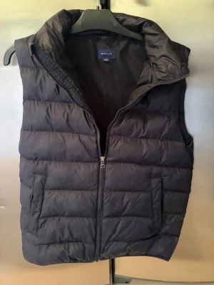 Svart dunväst från GANT - Snygg svart dunväst från GANT med dragkedja framtill och två fickor på sidorna. Västen har en hög krage och quiltad design som ger en sportig och modern look. Perfekt att slänga över en hoodie eller tröja för extra värme.