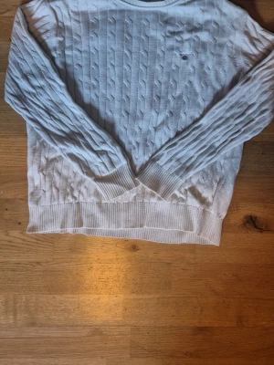 Vit kabelstickad tröja Gant - Vit kabelstickad tröja från Gant med rund halsringning och diskret logga på bröstet. Tröjan har långa ärmar och ribbade muddar vid ärmslut och nederkant. Perfekt för en clean och klassisk look.