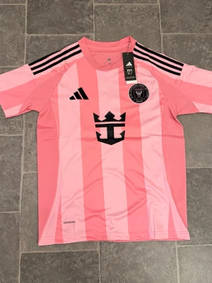 Inter Miami Messi #10 - Säljer en rosa Inter Miami matchtröja från Adidas med svarta detaljer och klubbmärke på bröstet. Tröjan har Messi och nummer 10 tryckt på ryggen samt svarta ränder på axlarna. Tillverkad i lätt och ventilerande material, perfekt för fotboll eller träning.