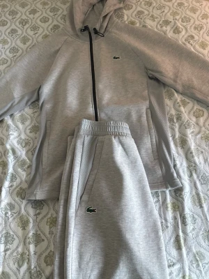 (Helt ny) Grå Lacoste tracksuit, tröja M, byxor S - Snygga gråa mjukisbyxor från Lacoste med klassisk krokodillogga på benet. Byxorna har resår i midjan och sidofickor. Perfekta för en avslappnad och sportig stil. Matchar med hoodie i samma färg och material.