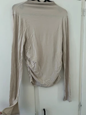Beige långärmad polotopp - Trendig beige polotopp med långa ärmar och rynkade sidor. Tillverkad i ett mjukt och stretchigt material som ger en slim passform. Perfekt att styla med jeans eller kjol för en clean look.