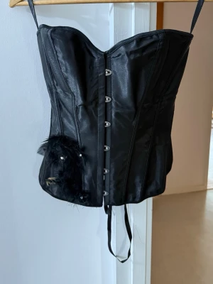 Svart korsett med fjädrar och snörning - Snygg svart korsett med avtagbara axelband, dekorativa fjädrar och blanka detaljer framtill. Stängs med hakar fram och har snörning bak för justerbar passform. Materialet ser ut som satin eller liknande tyg. Perfekt för dig som vill ha en edgy och unik look.