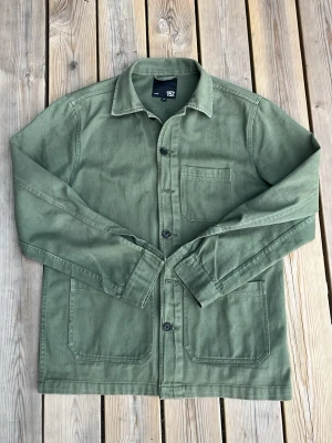 Olivgrön overshirt med fickor - Stilren olivgrön overshirt med tre stora utanpåfickor och knäppning framtill. Klassisk krage och raka ärmar. Tillverkad i ett robust bomullstyg som ger en avslappnad look. Perfekt att bära öppen över en t-shirt eller stängd som jacka. OBS Några knappar saknas därav priset!
