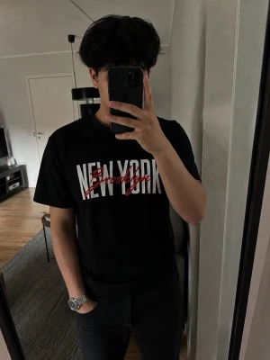 Svart t-shirt med New York-tryck - Snygg svart t-shirt med stort vitt tryck där det står 'NEW YORK' och röd text 'Brooklyn' över. Klassisk rund hals och korta ärmar. Perfekt för dig som gillar streetstyle och vill ha en enkel men cool look.