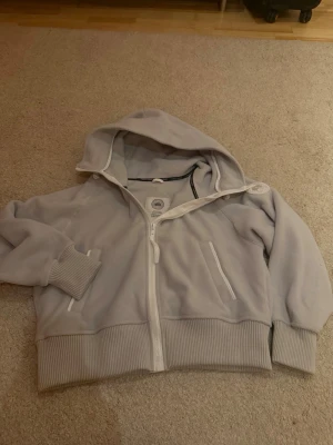Ljusgrå hoodie med dragkedja - Snygg ljusgrå hoodie med dragkedja framtill och två fickor. Hoodien har ribbade muddar vid ärmar och midja samt en stor huva med dragsko. Detaljer i vitt och en patch på ena ärmen ger en clean look. Perfekt för chill dagar eller när du vill vara extra bekväm.