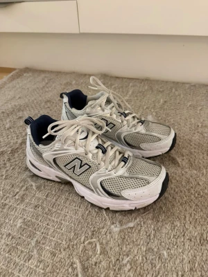New Balance 530 sneakers - Säljer ett par klassiska New Balance sneakers i vitt och silver med marinblå detaljer. Skorna har meshpaneler för extra ventilation, vita snören och en chunky sula. Perfekta för dig som gillar retrostil och sportig vibe. Knappt använda då de är för stora ! 