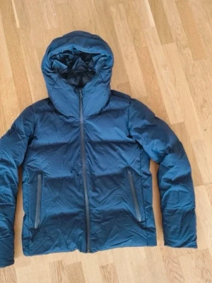 Blå pufferjacka med huva Uniqlo - Snygg blå pufferjacka från Uniqlo med stor huva och två dragkedjeförsedda fickor framtill. Jackan har en quiltad design och stängs med dragkedja. Perfekt för kalla dagar och ger en modern look. Materialet ser ut att vara syntetiskt och jackan är vadderad för extra värme.