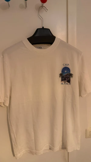 Vit t-shirt med Casa Blanca-tryck - Vit t-shirt med rund hals och korta ärmar. På bröstet finns ett färgglatt tryck med palmer och texten 'Casa Blanca' i blått. Perfekt för en avslappnad och stilren look.