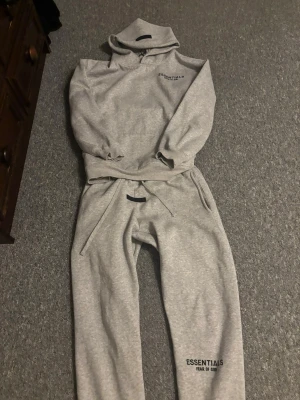 Grått Essentials set med huva - Säljer ett grått mjukisset från ESSENTIALS Fear of God. Setet består av en hoodie med huva och matchande byxor med dragsko och fickor. Båda delarna har logotyptryck. Perfekt för en chill och avslappnad stil.