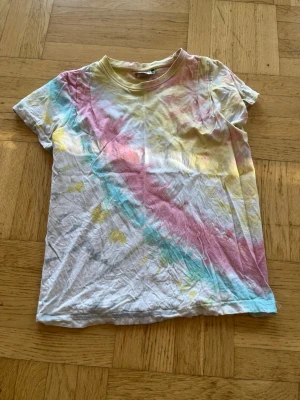 Färgglad tie-dye t-shirt från Clique - Säljer en färgglad tie-dye t-shirt från Clique i storlek XS. T-shirten har korta ärmar och är gjord i mjuk bomull. Färgerna går i pastellrosa, blått, gult och vitt, vilket ger en unik och somrig look. Perfekt för dig som gillar färg och vill sticka ut!