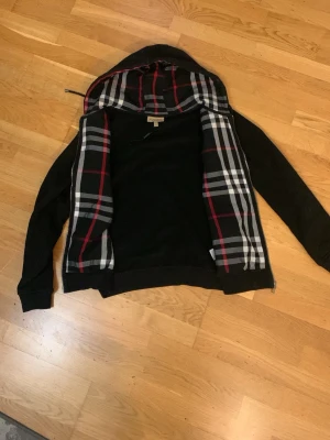 Svart hoodie med rutigt foder - Säljer en svart hoodie med dragkedja och huva. Insidan av huvan och öppningen har ett klassiskt rutigt mönster i svart, vitt och rött. Hoodien har långa ärmar och ribbade muddar. Perfekt för dig som gillar stilrena detaljer och vill ha något bekvämt.