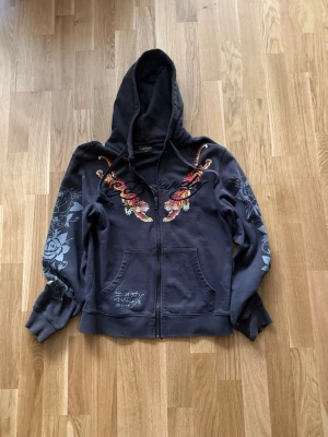 Svart hoodie med tigertryck Ed Hardy - Säljer en svart hoodie från Ed Hardy med dragkedja, huva och fickor framtill. Framsidan har broderade tigrar i orange, gult och vitt samt Ed Hardy-logga. Ärmarna har gråa blommönster. Perfekt för dig som gillar streetwear och detaljerade prints.