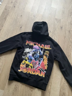 Svart Michael Jordan hoodie, Limited Edition - Säljer en svart hoodie med stor färgglad Michael Jordan-print på ryggen. Hoodien har huva och trycket visar flera bilder av Jordan i olika poser, samt hans namn i stora bokstäver. Perfekt för dig som gillar basket och streetwear. Från Iconic Family Member, Limited Edition, Clothing från STHLM