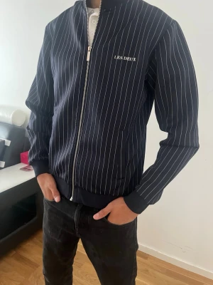 Mörkblå bomberjacka med vita ränder - Snygg mörkblå bomberjacka från Les Deux med tunna vita vertikala ränder. Jackan har dragkedja framtill, ribbade muddar vid ärmslut och nederkant samt två sidofickor. Tillverkad i ett mjukt och bekvämt material som ger en clean och modern look. 