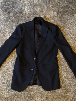 Mörkblå slim fit kavaj - Stilren mörkblå kavaj i slim fit-modell med två knappar framtill och tre knappar vid ärmslutet. Klassisk design med två fickor och bröstficka. Perfekt för dig som gillar en smalare passform och vill ha en tidlös look.