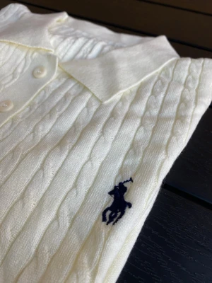 Vit kabelstickad polotopp - Kabelstickad vit polotopp med korta ärmar och krage. Toppen har tre knappar framtill och en diskret broderad logga på bröstet. Materialet känns mjukt och stretchigt, perfekt för en clean och klassisk look.
