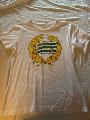 Vit HIF t-shirt med tryck - Hejsan 👋 jag säljer en vit t-shirt med stort HIF-tryck på bröstet. Trycket består av en grönvit flagga med bokstäverna HIF omgiven av en gul lagerkrans. Klassisk rund hals och korta ärmar. Perfekt för dig som vill visa ditt stöd för Hammarby. 💚🤍