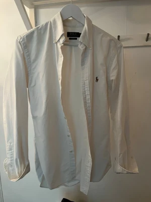 Vit Ralph Louren skjorta skjorta, bra skick 2 månader gammal.  - Klassisk vit skjorta med knappar framtill och button-down krage. Skjortan har lång ärm och en liten broderad logga på bröstet. Passar perfekt till jeans eller chinos för en clean och stilren look.