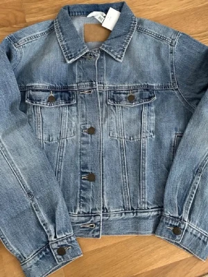 Klassisk blå jeansjacka M - Snygg blå jeansjacka i klassisk modell med raka sömmar och två bröstfickor med lock och knapp. Jackan har metallknappar framtill och vid ärmsluten. Perfekt att slänga över en hoodie eller t-shirt för en avslappnad look. Kort i längden cirka 27 cm från armhålan till nederkanten av tröjan