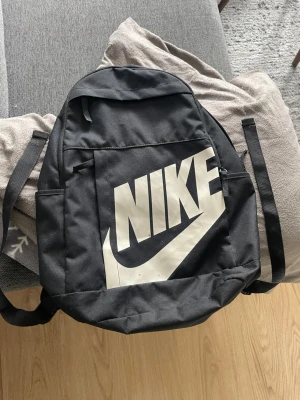 Svart ryggsäck från Nike - Snygg svart ryggsäck från Nike med stort vitt logotryck på framsidan. Ryggsäcken har justerbara, vadderade axelband och en stor ytterficka och de är 4 fickor 2 stora och 2 lite mindre med dragkedja. Tillverkad i slitstarkt tyg och har en klassisk, enkel form som passar till det mesta. Kan diskutera pris om de skulle vara relevant 